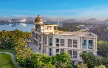 Elegant neoclassical-style building with a dome, surrounded by lush greenery and overlooking a serene lake with mountains in the background.