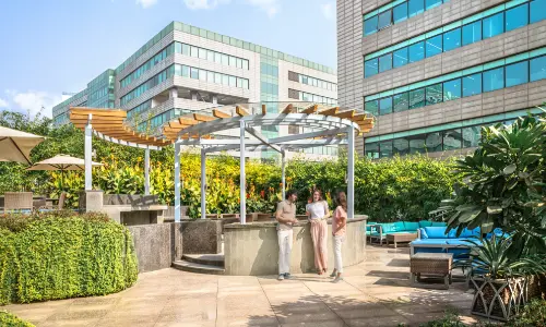 A modern outdoor lounge area with a pergola and comfortable seating at Candor TechSpace, Sector 48, Gurugram. Three people are enjoying the sunny atmosphere amidst greenery