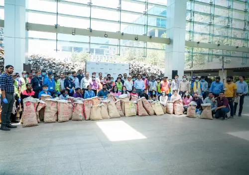 A group of individuals, dressed in work uniforms, proudly pose for a photo after completing a community cleanup drive. They are holding large bags filled with waste collected during the drive, with a large glass building in the background.