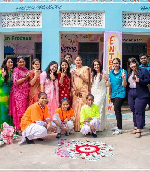 A group of people, including school students and staff, posing for a photo outside a school building with a colorful Rangoli design on the ground