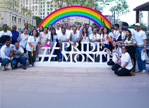 Group of employees posing together under a rainbow arch with the words #PrideMonth, celebrating diversity and LGBTQ+ inclusivity during Pride Month.