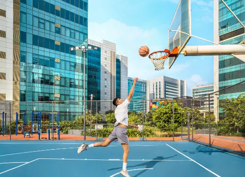 A man taking a jump shot at a basketball hoop in the workplace's outdoor sports area surrounded by modern office buildings.