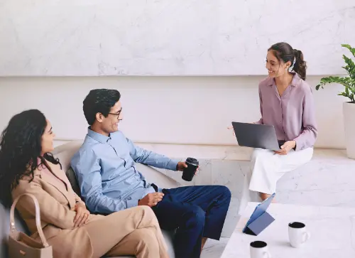 Three people in a relaxed meeting, with one woman sitting with a laptop while two individuals, one holding a coffee cup, listen and engage.