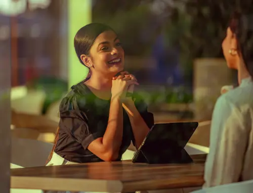 A woman smiling while sitting at a table and having a conversation with someone, with a tablet in front of her.