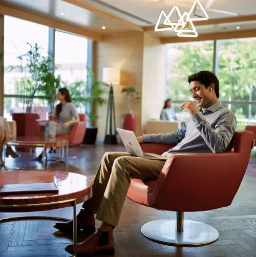 A person working on a laptop and smiling while seated in a comfortable chair at a modern office space with large windows.