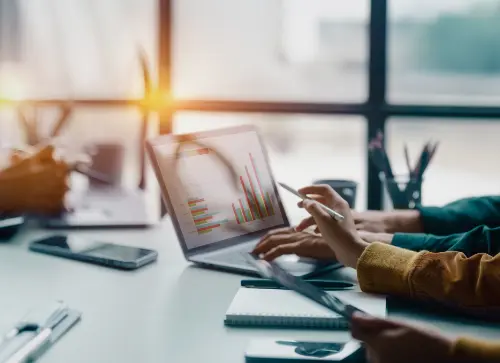 A close-up of a team reviewing data on a laptop, with graphs visible on the screen and individuals using pens and notebooks to discuss.