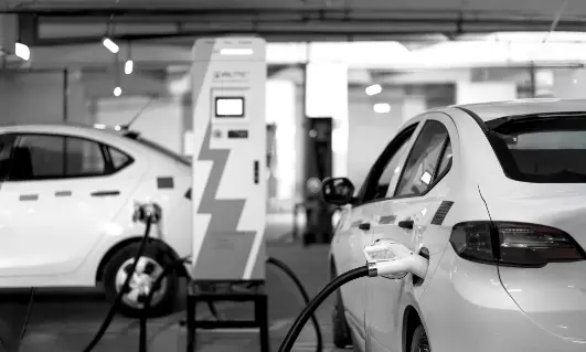 A black and white image of electric vehicles being charged at a charging station. The vehicles are connected to the charging point, which displays a lightning bolt icon, symbolizing electric energy. The setting is an indoor parking area with modern infrastructure for electric vehicle charging.