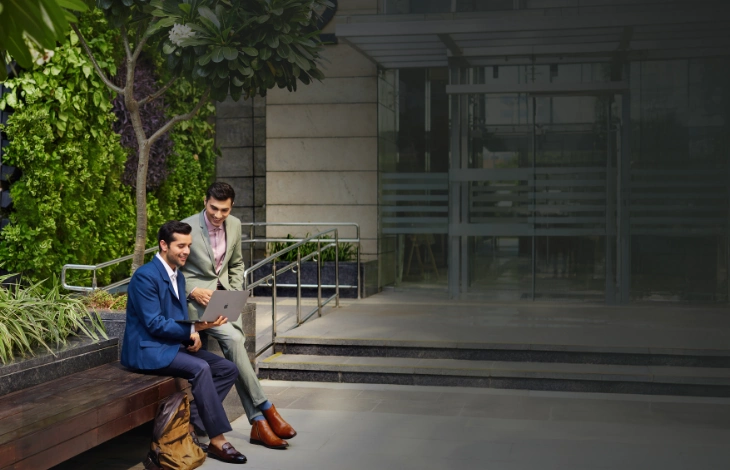 Two business professionals sit on a bench in a lush green courtyard at Candor TechSpace, Sector 135, Noida, collaborating on a laptop. The modern and serene environment enhances their productive discussion
