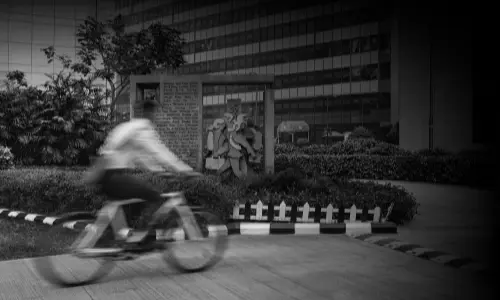 A black and white image of Candor TechSpace, Sector 48, Gurugram, featuring a blurred cyclist in motion in the foreground. In the background, there is a landscaped area with greenery, a small brick structure with a sculpture, and a modern glass office building.