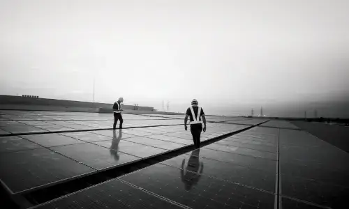 Two workers walking on a rooftop, inspecting solar panels. The image is in black and white, with the workers wearing safety gear and helmets
