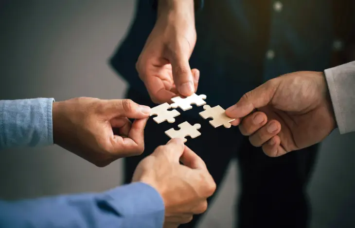 Four hands coming together to connect puzzle pieces, symbolizing teamwork and collaboration.