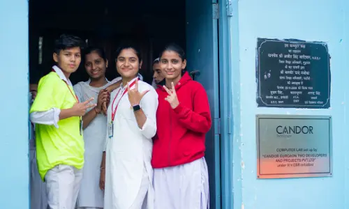  A group of smiling students standing at the entrance of a computer lab sponsored by Candor, making hand gestures and posing for the camera.