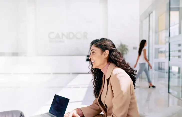 A woman smiling and engaging in a conversation with a colleague at Candor TechSpace, Sector 135, Noida. The modern office space is bright and spacious with glass walls, and another person is seen walking in the background