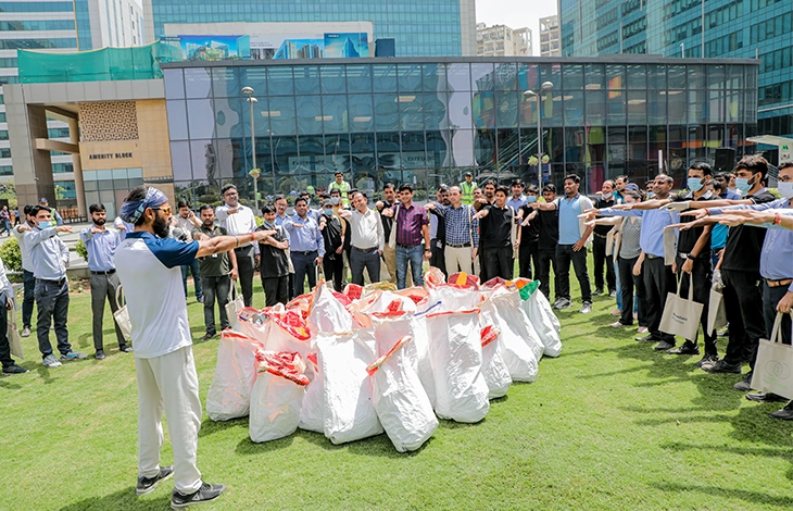 A group of people standing in a circle outdoors, participating in an event, with a leader giving instructions as they prepare to collect trash in large bags