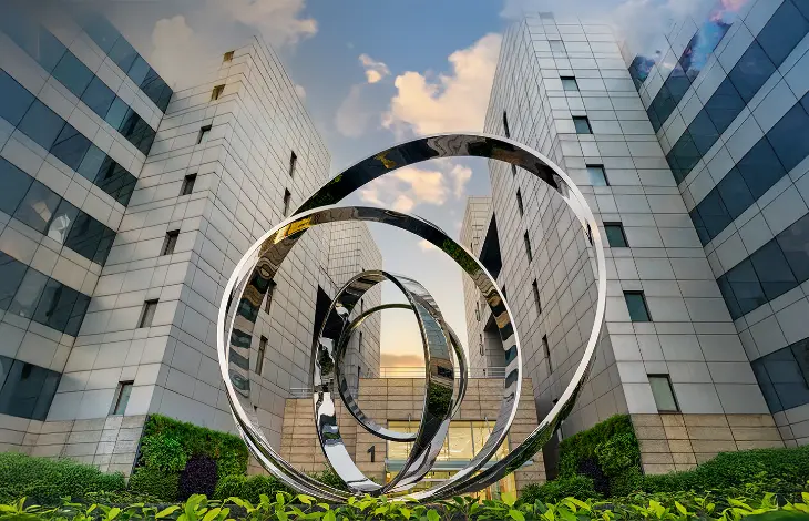 A modern office complex at Candor TechSpace, Sector 135, Noida, featuring a striking metallic circular sculpture in the landscaped courtyard.