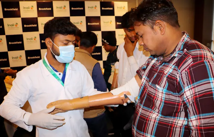 A medical professional administering a blood test to a person at a health camp, with people and a Brookfield backdrop in the background