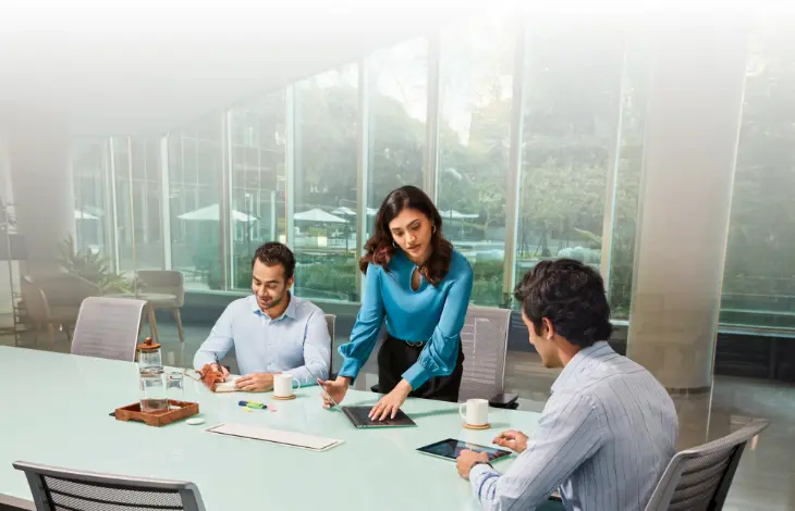 A team of three professionals in a bright and modern office meeting room. A woman is writing on a tablet, while two men are engaged with a notebook and a tablet in front of them. They are collaborating around a glass table with a clear view of the outdoors in the background.