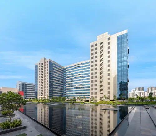  A modern office building at Candor TechSpace, Sector 21, Gurugram, reflecting in a water body with clear blue skies above.