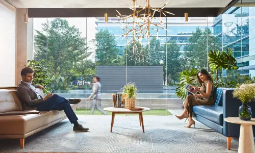 A modern office lounge with comfortable seating, featuring two individuals—one reading on a sofa and the other seated on a blue chair, both in a well-lit space with glass walls overlooking lush greenery. A person can be seen walking outside the window