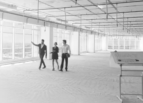 Three professionals, two men and one woman, walk through an empty office space with exposed ceilings and large windows, discussing the layout or potential design for the space.