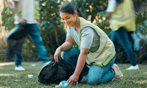A woman wearing a safety vest, smiling as she kneels on the grass to pick up litter, part of a group working together for environmental cleanup.
