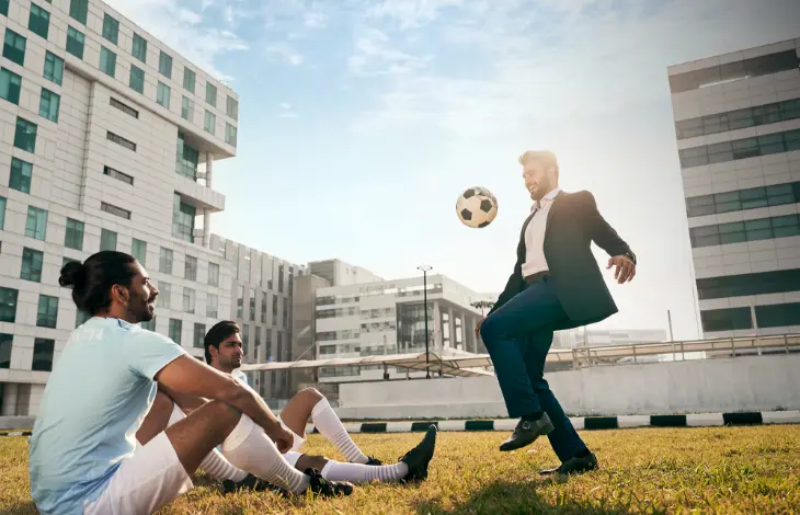 A man in a suit playing football with two others in sportswear at Candor TechSpace, Newtown, Kolkata, with modern buildings in the background.