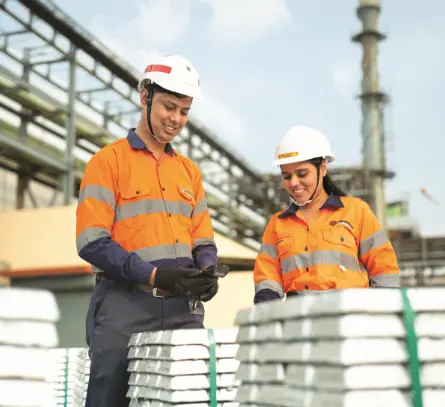 Two Hindustan Zinc employees in uniforms inspecting stacked zinc ingots at an industrial site.