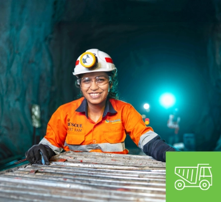 Female mining professional wearing an orange safety uniform and helmet underground, with safety lights illuminating the tunnel.