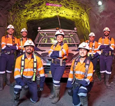 Group of miners wearing safety gear and orange uniforms underground mine with a mining vehicle.