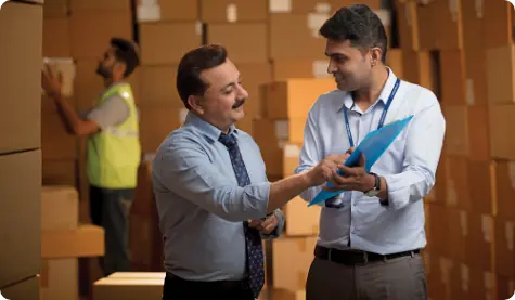 Two professionals engaged in a discussion inside a warehouse, surrounded by stacked cardboard boxes. One of them, holding a blue folder, is explaining something to the other, who is listening attentively. In the background, a worker is organizing the boxes. The image depicts a typical business or logistics environment focused on inventory management.