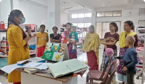 A child nutritionist conducts a counselling session at Cachar Cancer Care Hospital and Research Centre in Silchar, Assam. The session involves several women and children gathered around a table, discussing nutrition and health. The room is well-lit with large windows in the background, and educational materials are displayed on the table to support the session.