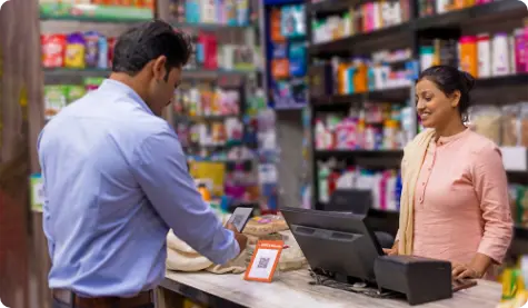 A scene in a local store where a customer is making a digital payment using a mobile device. The store cashier, a woman in traditional attire, smiles while overseeing the transaction. The image highlights the growing use of digital payment systems in everyday retail settings.